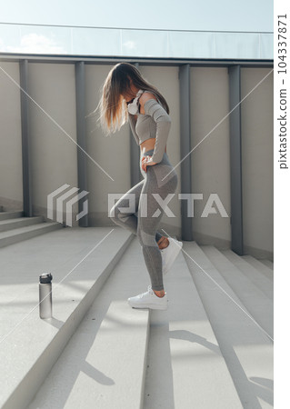 Sporty woman doing jumps and squats on stairs. Wearing grey sports active wear and headphones 104337871