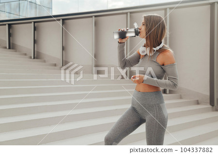 Fitness woman in gray tracksuit drinking water outdoors after workout Fitness woman in gray tracksuit drinking water outdoors after workout 104337882