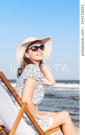 Happy brunette woman wearing sunglasses while relaxing on a wooden deck chair at the ocean beach 104338081