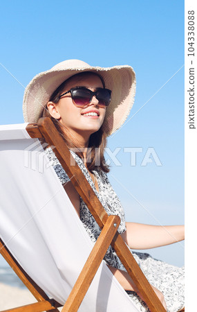 Happy brunette woman wearing sunglasses and hat relaxing on a wooden deck chair at the ocean beach Happy brunette woman wearing sunglasses and hat relaxing on a wooden deck chair at the ocean beach 104338088