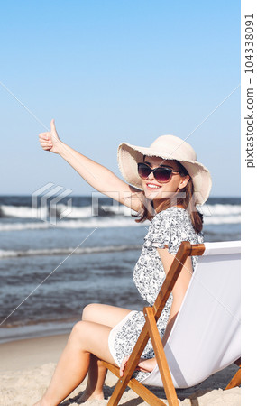 Happy brunette woman sitting on a wooden deck chair at the ocean beach while showing Thumb up Happy brunette woman sitting on a wooden deck chair at the ocean beach while showing Thumb up 104338091