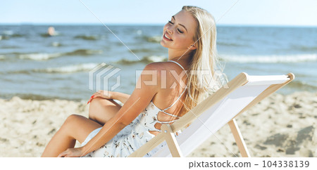 Happy blonde woman relaxing on a wooden deck chair at the ocean beach Happy blonde woman relaxing on a wooden deck chair at the ocean beach 104338139