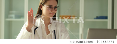 Woman doctor in glasses waving hand at laptop screen in clinic Woman doctor in glasses waving hand at laptop screen in clinic 104338274