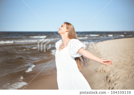 Happy smiling beautiful woman is on the ocean beach in a white summer dress, open arms 104339139