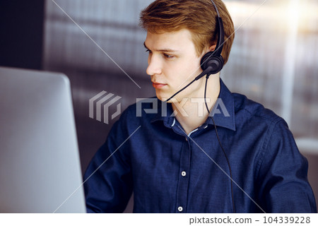 Young blond businessman using headset and computer in a darkened office, glare of light on the background. Startup business means working hard Young blond businessman using headset and computer in a darkened office, glare of light on the background. Startup business means working hard 104339228