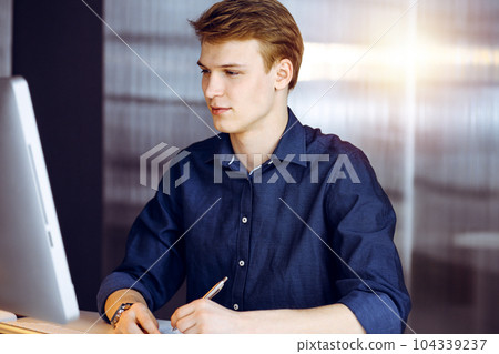 Young blond businessman thinking about strategy at his working place with computer in a darkened office, glare of light on the background Young blond businessman thinking about strategy at his working place with computer in a darkened office, glare of light on the background 104339237