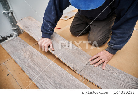 Close up of male hands laying laminate wooden planks on the floor in apartment under renovation. Man construction worker installing timber laminate flooring at home. Hardwood floor renovation concept. 104339255