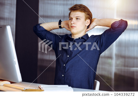 Young blond businessman and programmer stretching hands after working hard with computer in a darkened office, glare of light on the background 104339272