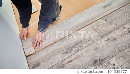 Close up of man construction worker installing laminate wooden flooring in apartment under renovation. Male hands placing laminate timber board on the floor at home. Flooring renovation concept. Close up of man construction worker installing laminate wooden flooring in apartment under renovation. Male hands placing laminate timber board on the floor at home. Flooring renovation concept. 104339277