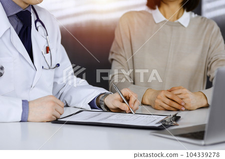 Unknown male doctor and patient woman discussing something while sittingin a darkened clinic and using clipboard, glare of light on the background Unknown male doctor and patient woman discussing something while sittingin a darkened clinic and using clipboard, glare of light on the background 104339278