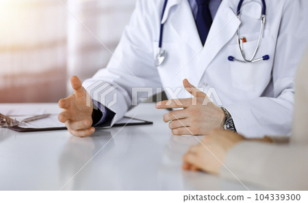 Unknown male doctor and patient woman discussing something while sittingin a darkened clinic, glare of light on the background. Close-up of hands Unknown male doctor and patient woman discussing something while sittingin a darkened clinic, glare of light on the background. Close-up of hands 104339300