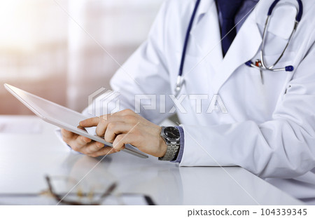 Unknown male doctor sitting and working with tablet computer iin a darkened clinic, glare of light on the background, close-up of hands 104339345