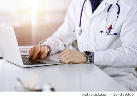 Unknown male doctor sitting and working with laptop in a darkened clinic, glare of light on the background, close-upof hands 104339360