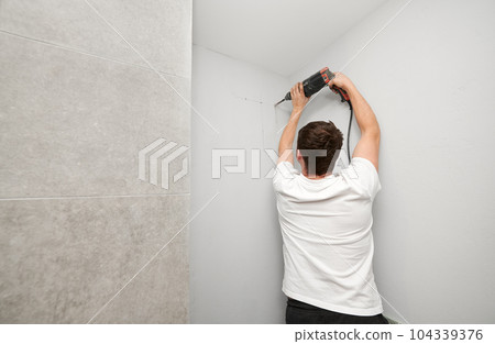 Back view of male person in white shirt using electric drilling machine while working on home renovation. Man drilling white wall with power drill tool before plumbing works in bathroom at home. 104339376
