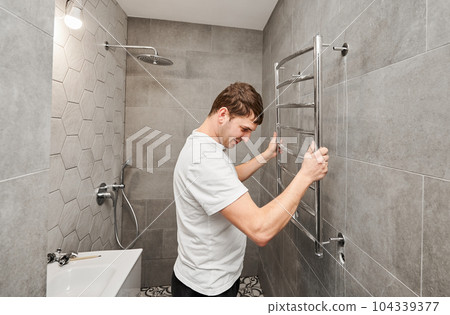Plumber hanging electric heated towel rack on the wall in bathroom. Man installing stainless steel bath towel heater in apartment under renovation. Home renovation concept. 104339377