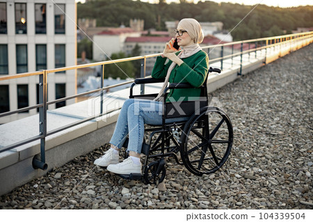 Lady in hijab chatting via gadget in wheelchair outside home Lady in hijab chatting via gadget in wheelchair outside home 104339504