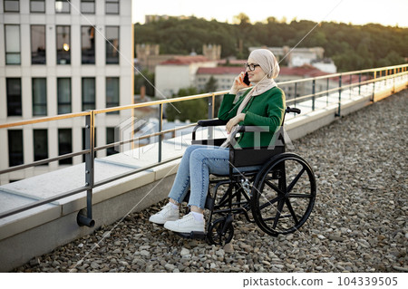 Lady in hijab chatting via gadget in wheelchair outside home 104339505
