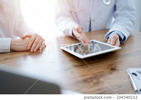Doctor and patient sitting at the wooden desk in clinic. Female physician's hands pointing into laptop computer monitor, close up. Medicine concept 104340152