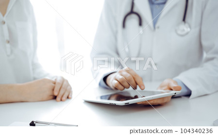 Doctor and patient sitting near each other at the desk in clinic. The focus is on female physician's hands pointing into tablet computer touchpad, close up. Medicine concept 104340236