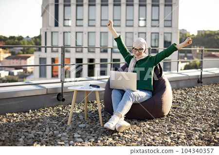 Female in hijab raising arms on rooftop with computer 104340357
