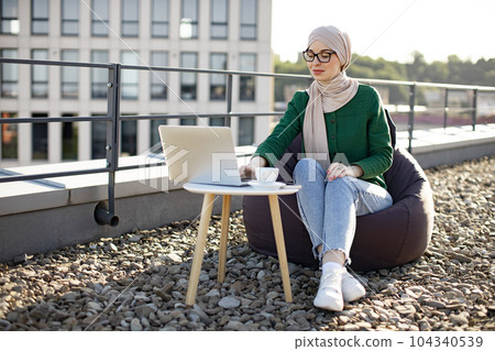 Lady in headscarf running laptop in beanbag on roof terrace Lady in headscarf running laptop in beanbag on roof terrace 104340539