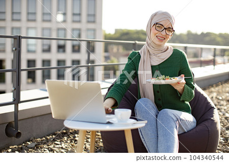 Lady holding plate with food while typing on laptop outside 104340544