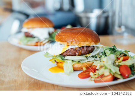 Closeup homemade burger with artificial meat, fried egg and vegetables on white plate with fresh salad on the kitchen table. Healthy fast food cooking recipe at home. Vegetarian diet. Selective focus. Closeup homemade burger with artificial meat, fried egg and vegetables on white plate with fresh salad on the kitchen table. Healthy fast food cooking recipe at home. Vegetarian diet. Selective focus. 104342192