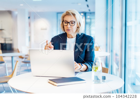 50's confident mature businesswoman having a video call chat on laptop. Middle-aged experienced senior female professional working on laptop in open space office. Female entrepreneur working remotely 50's confident mature businesswoman having a video call chat on laptop. Middle-aged experienced senior female professional working on laptop in open space office. Female entrepreneur working remotely 104342228