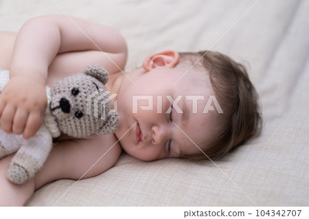 Sweet little boy with dark hair lying on white sheet in crib holding knitted bear toy with hand innocent chubby baby with closed eyes sleeping in nursery at home in early morning Sweet little boy with dark hair lying on white sheet in crib holding knitted bear toy with hand innocent chubby baby with closed eyes sleeping in nursery at home in early morning 104342707