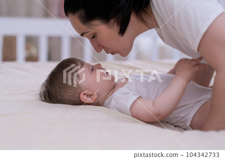 Loving mom leaning to kiss adorable little son lying on soft bed in nursery at home happy mother enjoying spending morning with chubby baby boy in white bodysuit 104342733