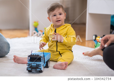 Cute chubby boy in yellow jumpsuit playing with blue truck toy sitting on white fluffy rug near mother woman spending time with little son in nursery at home 104342734