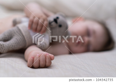 Cute baby boy lying on soft bed putting hand with tiny fingers on white sheet sweet chubby kid hugging knitted bear toy sleeping in nursery at home closeup 104342735