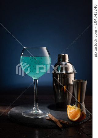 Bar composition on the table. Wine glass with a bright turquoise drink and an iron shaker. Orange slices and cinnamon sticks. Bar composition on the table. Wine glass with a bright turquoise drink and an iron shaker. Orange slices and cinnamon sticks. 104343240