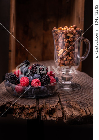 Various berries in a glass plate. Chocolate granola in a glass. Composition on the old board and on the background of the box. Various berries in a glass plate. Chocolate granola in a glass. Composition on the old board and on the background of the box. 104343285