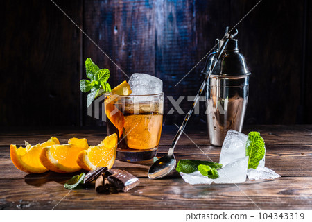A glass of whiskey and a large ice cube. Steel shaker and bartender's spoon. Sliced citrus and mint. A glass of whiskey and a large ice cube. Steel shaker and bartender's spoon. Sliced citrus and mint. 104343319