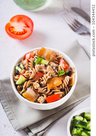 Fresh salad of buckwheat pasta, tomatoes, cucumbers and fried bacon in a bowl vertical view 104344452