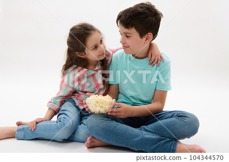 Adorable nice kid girl in plaid shirt and casual denim, gently hugging her older brother serving her a bowl of delicious popcorn while film screening, isolated on white studio background. Happy family 104344570