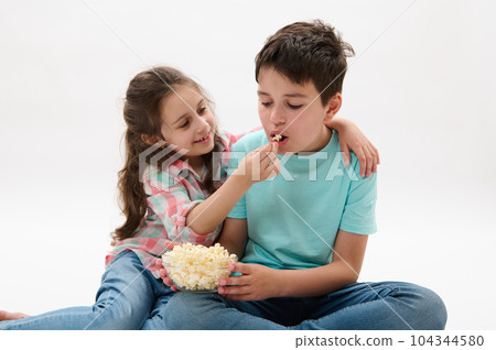 Lovely little girl hugging and feeding her older brother with tasty popcorn while watching movie or cartoons, isolated on white studio background. Kids. Entertainment. Leisure. Family relationships 104344580