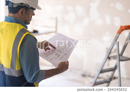 Concentrated worker in helmet checking apartment drawing stands in room in process of renovation experienced specialist in uniform standing in room against ladder near shabby wall 104345652