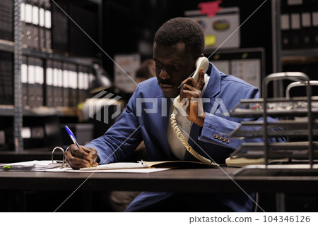African american police officer talking with remote detective using landline phone, discussing confidential clue. Private inspector analyzing crime scene evidence trying to solve case in arhive room 104346126