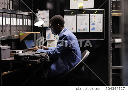 Bookkeeper reading administrative report, organizing management documents in storage room. African american businessman wearing formal suit working late at night in corporate depository 104346127