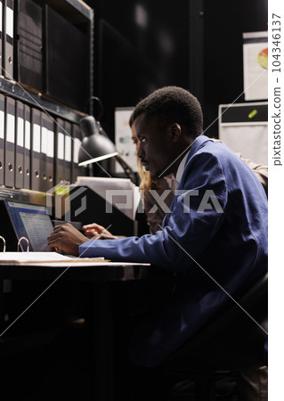 Diverse team working overtime at criminal case, analyzing crime scene evidence in arhive room. Private detectives searching new clues trying to catch mysterious suspect. Criminology concept 104346137