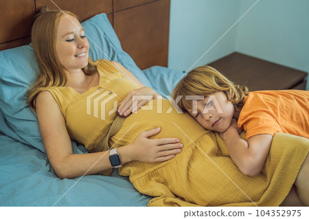 Young pregnant mother hugging her little boy lying in bed at night. Caring and loving parent hugging child 104352975