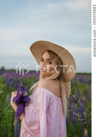 A beautiful woman in a straw hat walks in a field with purple flowers. A walk in nature in the lupin field A beautiful woman in a straw hat walks in a field with purple flowers. A walk in nature in the lupin field 104354883