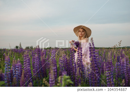 A beautiful woman in a straw hat walks in a field with purple flowers. A walk in nature in the lupin field 104354887