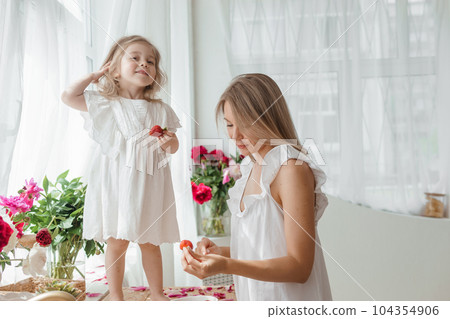 A little blonde girl with her mom on a kitchen countertop decorated with peonies. The concept of the relationship between mother and daughter. Spring atmosphere. A little blonde girl with her mom on a kitchen countertop decorated with peonies. The concept of the relationship between mother and daughter. Spring atmosphere. 104354906