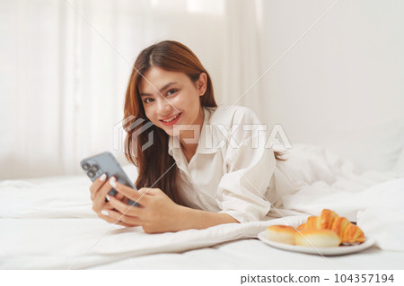 Happy smiling asian woman stretching on bed in the morning and holding mobile phone 104357194