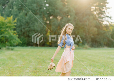 Young Caucasian woman or girl in a summer dress and a denim shirt is walking on the grass, holding shoes in her hands, relaxing outside the city on a weekend in summer 104358529