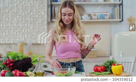 Young girl adds fresh chopped tomatoes to salad. Woman in home kitchen preparing healthy vegetable salad smiles enjoy process. Healthy eating and cooking concept. Young girl adds fresh chopped tomatoes to salad. Woman in home kitchen preparing healthy vegetable salad smiles enjoy process. Healthy eating and cooking concept. 104359012