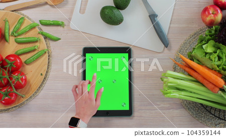 Woman using a tabletPC with a green screen at the kitchen table with fresh vegetables and fruits. Application for cooking. Healthy food concept. Woman using a tabletPC with a green screen at the kitchen table with fresh vegetables and fruits. Application for cooking. Healthy food concept. 104359144